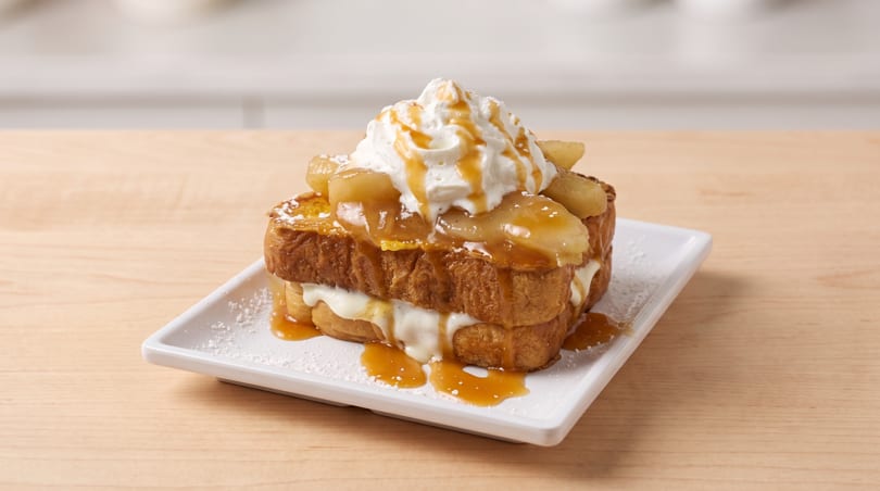 Stacked French toast with whipped cream, caramelized apples, caramel drizzle, and powdered sugar on a white plate.