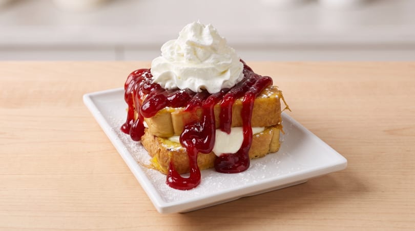 Two pieces of french toast topped with whipped cream, cherry sauce, and powdered sugar on a white plate.